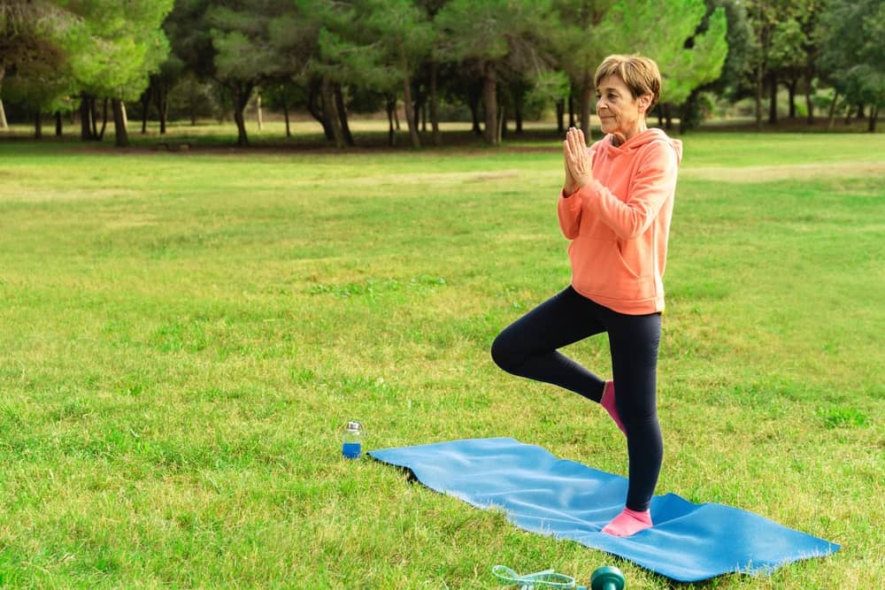 A senior woman balances on one leg on a yoga matt outside
