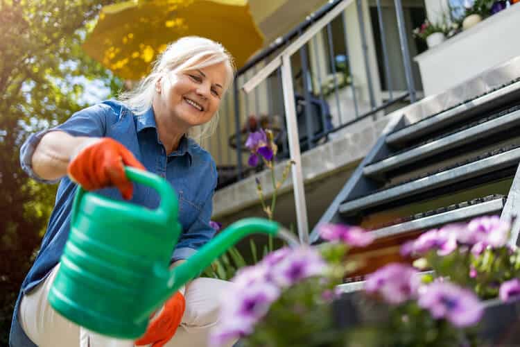 A senior woman waters flowers in her garden for health benefits.